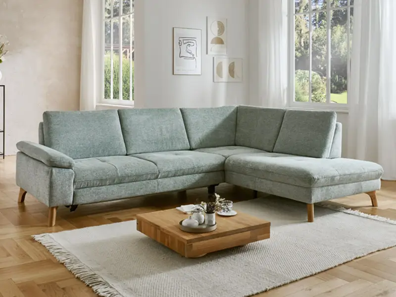 Helles Wohnzimmer mit einer hellgrauen, modernen Eckcouch, kleinem hölzernen Couchtisch auf einem beigen Teppich und großen Fenstern im Hintergrund.