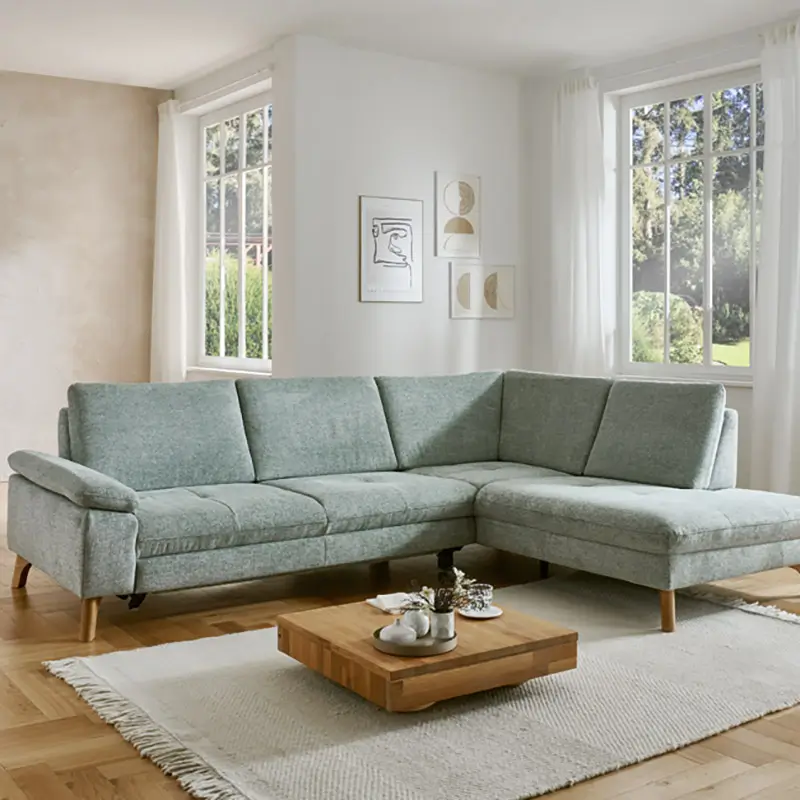 Hellgrünes, modernes Ecksofa mit Holzbeinen in einem hellen Wohnzimmer mit Holzboden, zwei Fenstern und minimalistischer Wanddekoration.