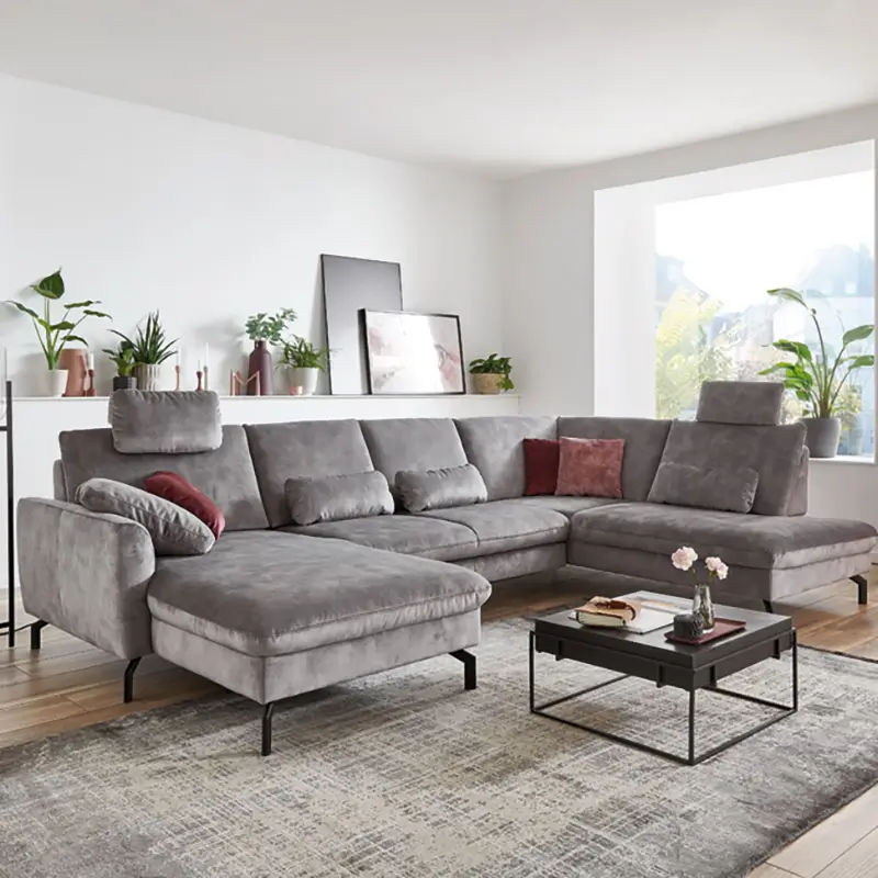 Großes modernes graues Ecksofa mit Kissen und Kopfstützen in einem hellen Wohnzimmer mit Pflanzen und Couchtisch.