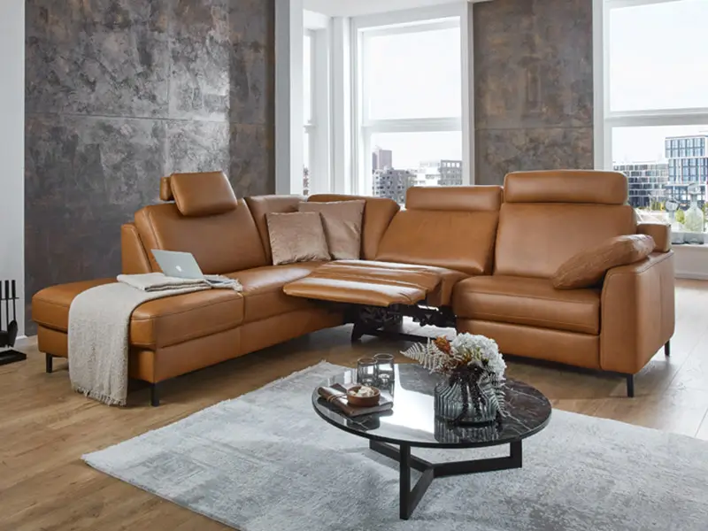 Modernes braunes Ledersofa mit verstellbarer Fußstütze und Kopfstützen in einem hellen Wohnzimmer mit großem Fenster.