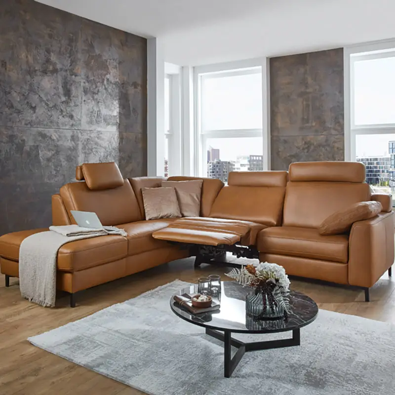 Moderner Raum mit braunem Ledersofa mit ausklappbarer Fußstütze, hellgrauem Teppich und einem schwarzen Glastisch mit Blumenstrauß.