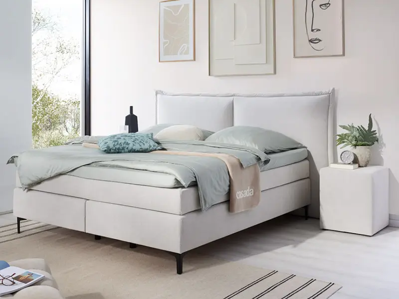 Modernes Boxspringbett in hellem Grau mit gepolstertem Kopfteil in einem minimalistisch eingerichteten Schlafzimmer.