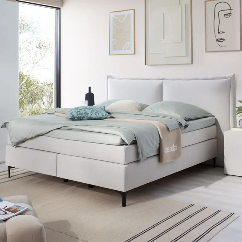 Modernes hellgraues Boxspringbett mit hellblauer Bettwäsche, Kissen und dekorativer Decke in einem hellen Schlafzimmer mit Fenster und minimalistischer Wandkunst.
