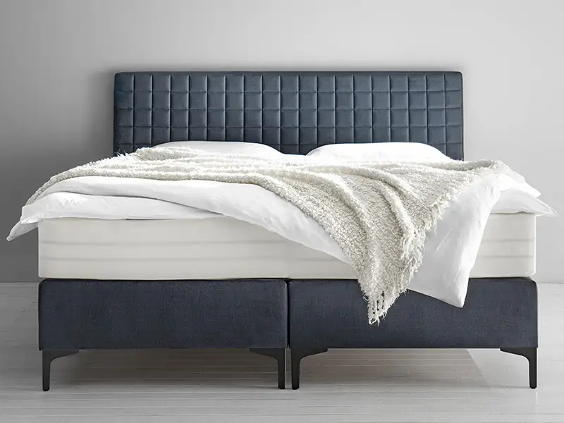 Modernes Boxspringbett mit dunkelgrauem Kopfteil und Untergestell, weißer Bettwäsche und cremefarbenem Überwurf in minimalistischem Schlafzimmer.