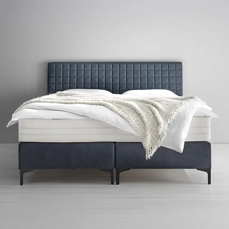 Modernes Boxspringbett mit dunkelgrauem gepolstertem Kopfteil, weißer Matratze, Bettdecke und grauer Kuscheldecke.