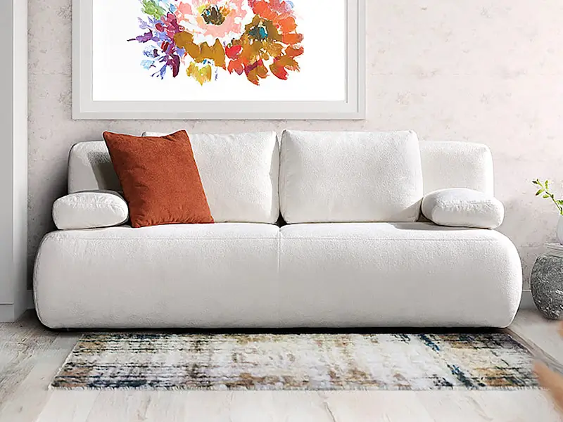 Weißes Sofa mit zwei runden Armlehnen und einem orangefarbenen Kissen vor einer Wand mit buntem Blumenbild und einem Teppich auf dem Boden.