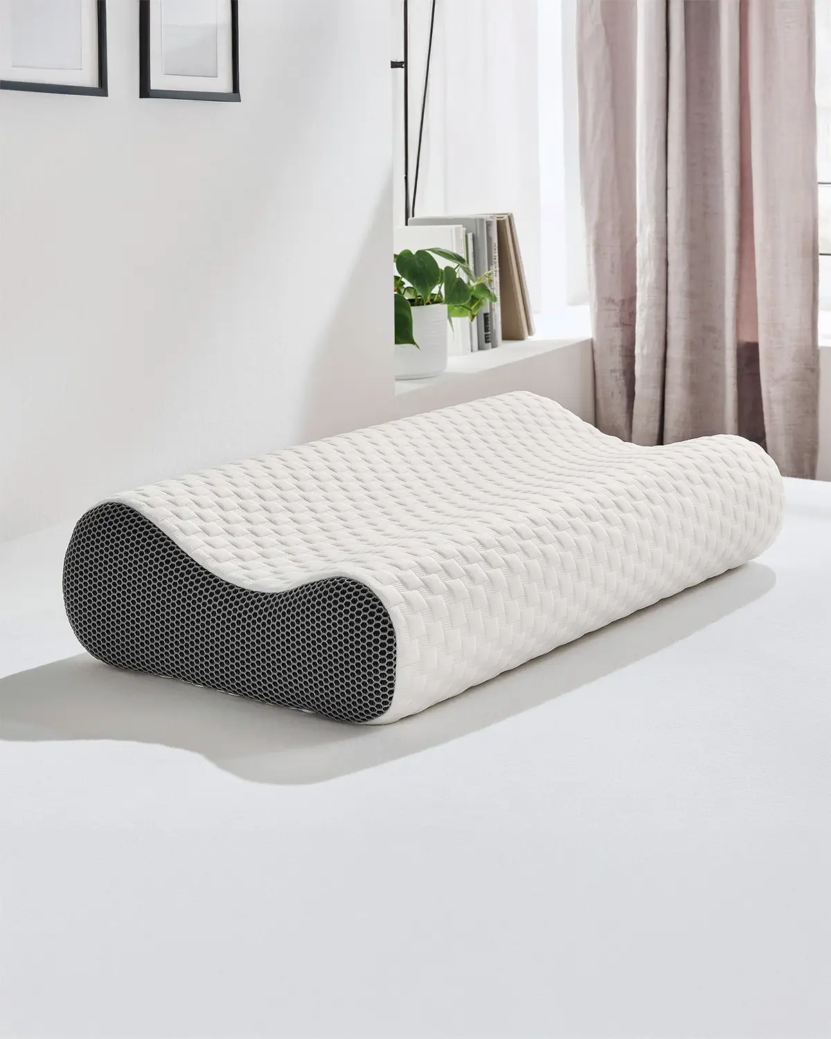 Orthopädisches Nackenstützkissen mit weißem, gewebtem Bezug auf einer weißen Oberfläche in einem hellen Raum mit Pflanzen und Vorhängen.